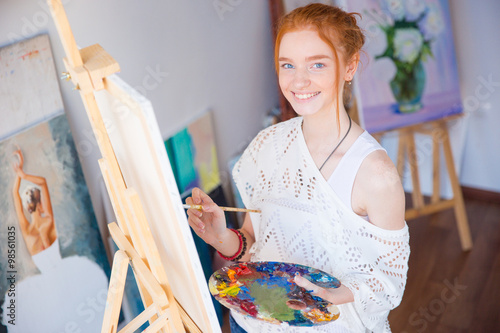 Cheerful woman artist standing and painting picture in workshop
