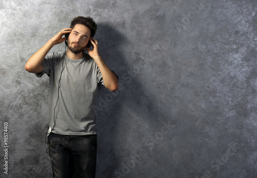 Young man listening music with headphones on grey wall background