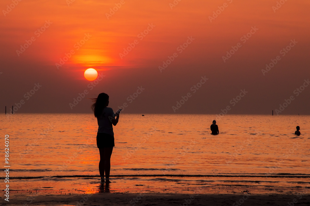 Silhouette of woman holding smart phone, standing on the beach at sunset