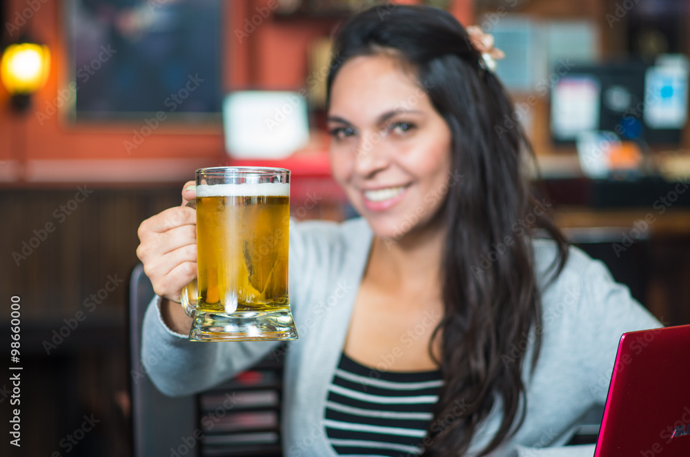 Brunette model sitting by restaurant table holding glass of beer and posing with positive attitude smiling Brunette model sitting by restaurant table holding glass of beer and posing with positive attitude smiling