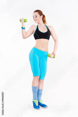 Young athletic red-haired woman in sportswear doing exercise with dumbbells. She smiles brightly.