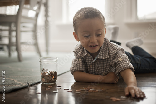 A child lying on his stomach on the floor playing with coins and putting them in a glass jar.  photo