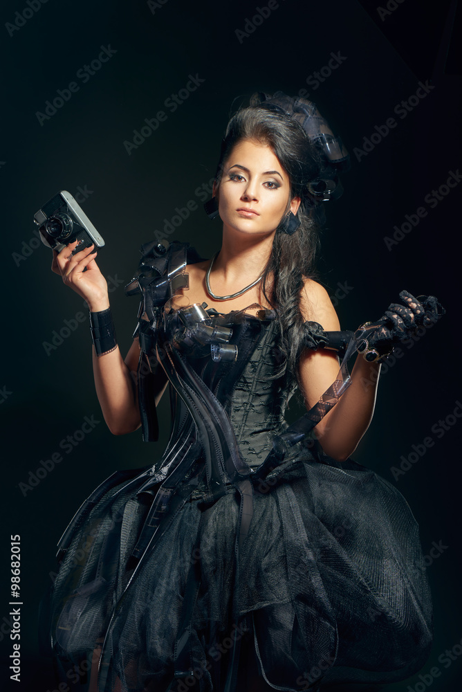 Beautiful young brunette woman in a black dress decorated with film camera rolls holding retro camera Beautiful young brunette woman in a black dress decorated with film camera rolls holding retro camera