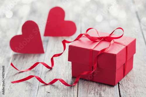 Gift box with red bow ribbon and two paper heart on wooden background for Valentines day