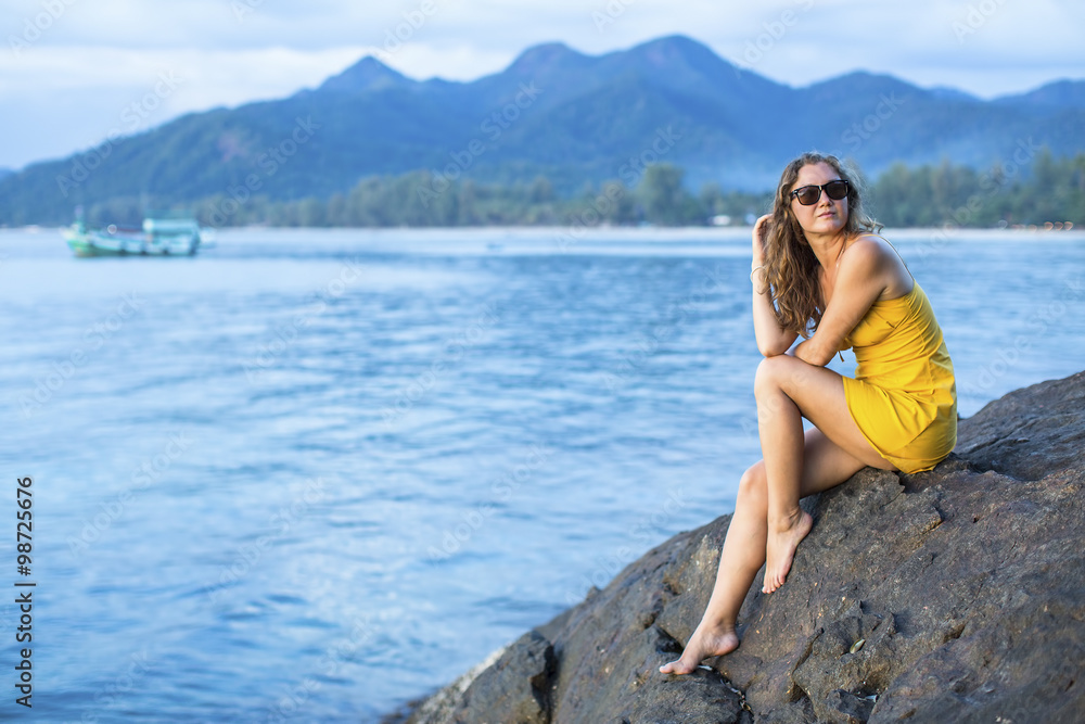 Beautiful young woman in a yellow dress, sits on a cliff overlooking the Sea in the evening time.