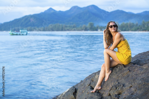 Beautiful young woman in a yellow dress, sits on a cliff overlooking the Sea in the evening time. © De Visu