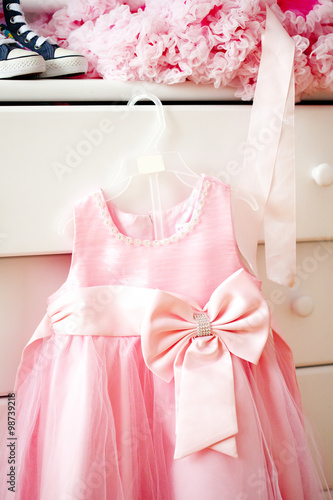 Розовое платье для девочки висит на комоде с вещами: кедами, юбочками, бусами и другими аксессуарами