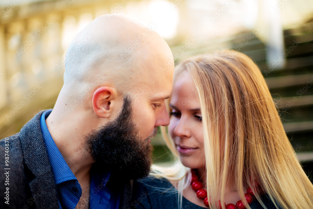 Fototapeta premium One beautiful stylish couple of young woman and senior man with long black beard sitting embracing close to each other outdoor in autumn street on stairs sunny day, horizontal picture