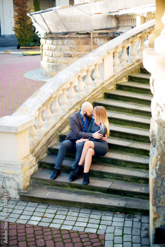 One beautiful stylish couple of young woman and senior man with long black beard sitting embracing close to each other outdoor in autumn street on stairs sunny day, horizontal picture