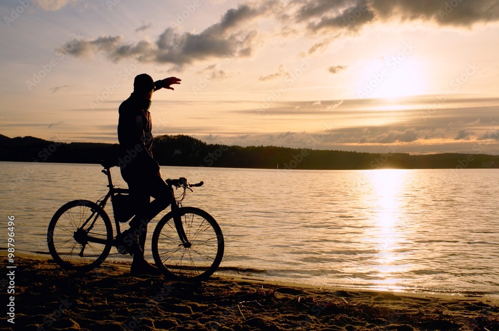 Silhouette of sportsman  holding bicycle on lake bech, colorful  sunset cloudy sky and reflection in wavy water level