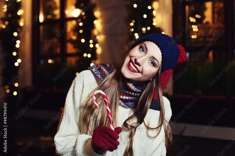Night street portrait of smiling  beautiful young woman with Christmas candy cane. Model looking at camera. Lady wearing classic winter knitted clothes. Festive garland lights. 