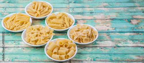 Various dried pasta variety and shapes in white bowl over wooden background photo