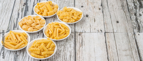 Various dried pasta variety and shapes in white bowl over wooden background