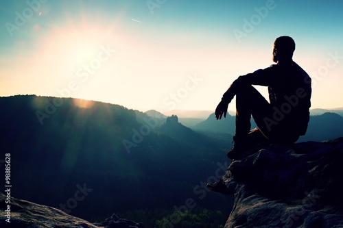 Tourist take a rest. Handsome young man sitting on the rock and enjoying view into misty rocky mountains.