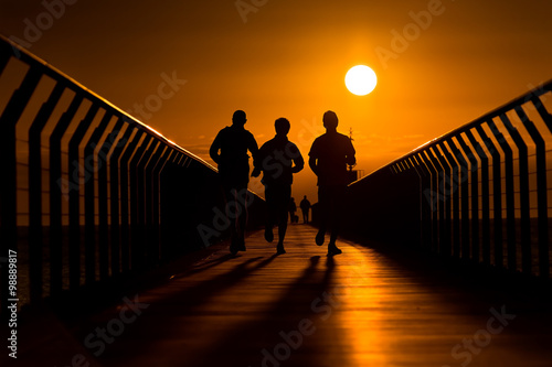 Silhouette of three backlit runners running towards the rising sun at the end of pier.