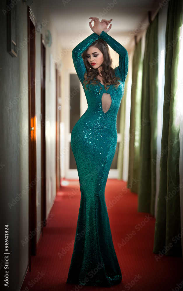 Young beautiful luxurious woman in long elegant turquoise dress posing  indoors. Attractive brunette with tight fit glamorous dress in hotel lobby.  Fashionable seductive female on red carpet of hall Stock-Foto | Adobe