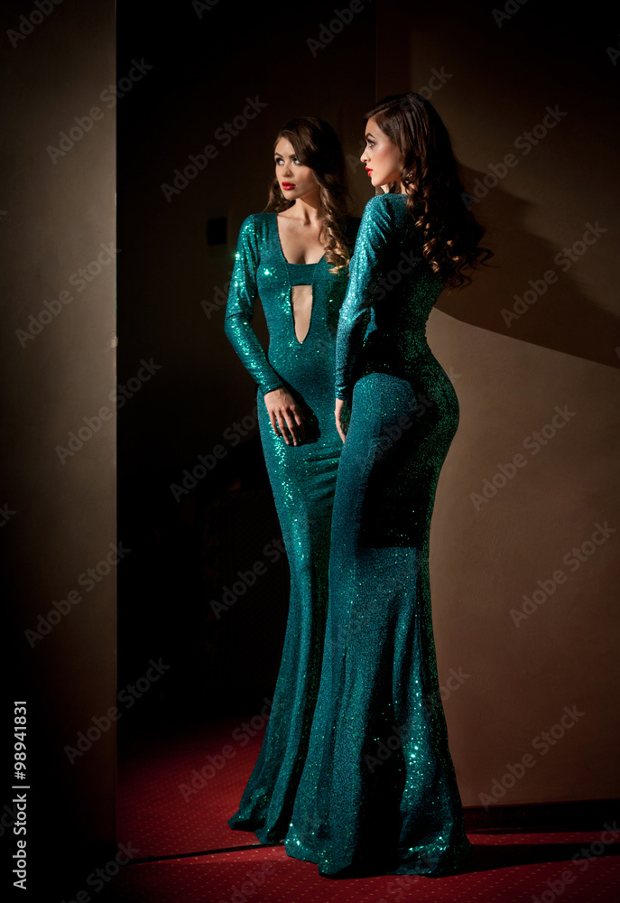 Elegant young woman in turquoise long dress looking into a large mirror, side view. Beautiful slim girl with creative hairstyle posing in front of a wall mirror, indoors shot. Fashionable model.