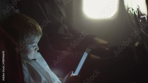 Young boy is playing with tablet on an airplane while father talks to him next to a window.