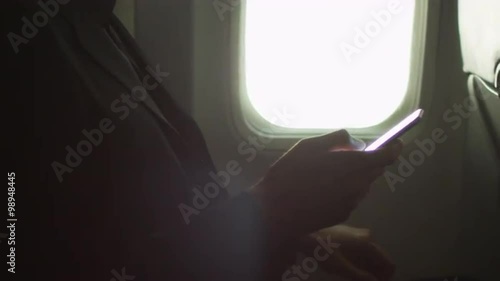 Young man in close-up is using a smartphone inside an airplane next to a window.