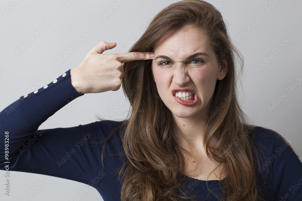 Obraz premium despair and humor concept - aggressive 20s girl pretending killing herself,grinding teeth with finger pointing to her temple,studio shot