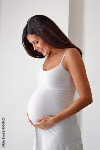 Feliz embarazada en casa tocando barriga con vestido blanco.