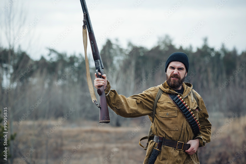 Fototapeta premium brutal hunter, bearded man in warm hat with a gun in his hand, a knife a backpack and smoking pipe in the wild forest in the autumn
