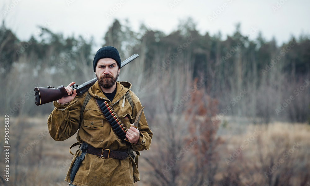 Fototapeta premium brutal hunter, bearded man in warm hat with a gun in his hand, a knife a backpack and smoking pipe in the wild forest in the autumn