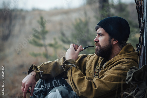 brutal hunter, bearded man in warm hat with a gun in his hand, a knife a backpack and smoking pipe in the wild forest in the autumn