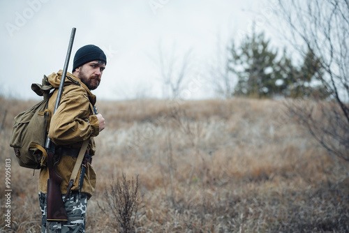 Fotografiet brutal hunter, bearded man in warm hat with a gun in his hand, a knife a backpac