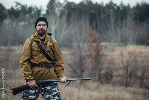 Photography brutal hunter, bearded man in warm hat with a gun in his hand, a knife a backpac