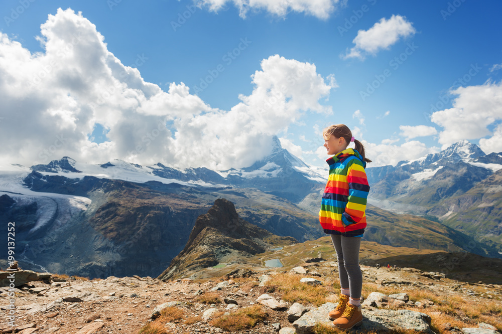 Cute little girl wearing bright rainbow colored coat and beige boots, resting in mountains,  Switzerland