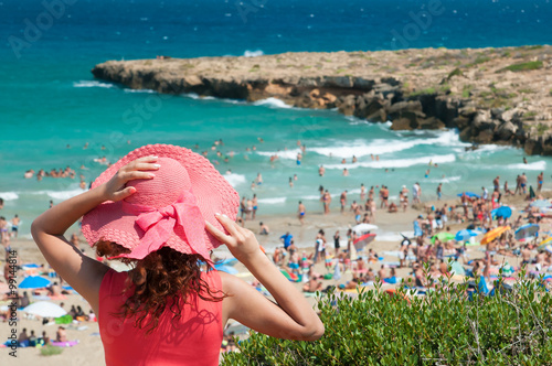 Tourist girl with a pink hat looking at the crowded beach of Calamosche, Sicily, from a panoramic point photo