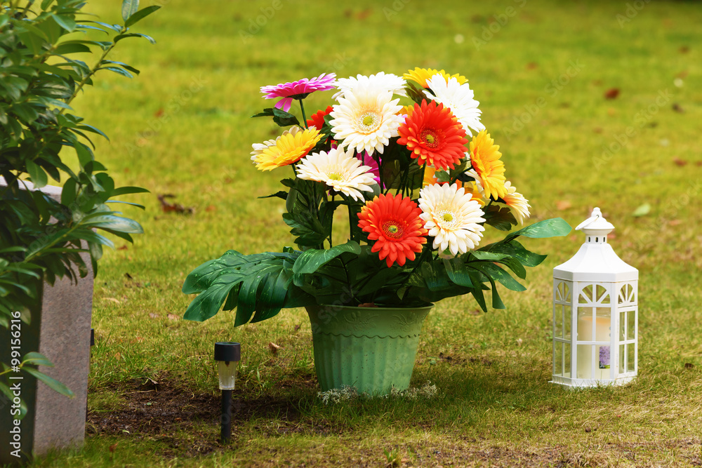 Colorful plastic flower in a pot beside an unknown headstone. A small white  lantern with candle inside stand beside pot. Smaller black solar powered  lamp stuck in the ground as well. Stock