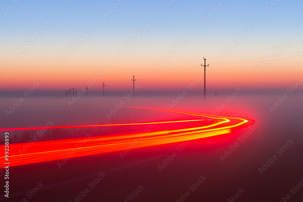 Long Exposure Red Car light trails on a road outside at foggy night on blue hour with electrical power lines and pylons disappear over the horizon