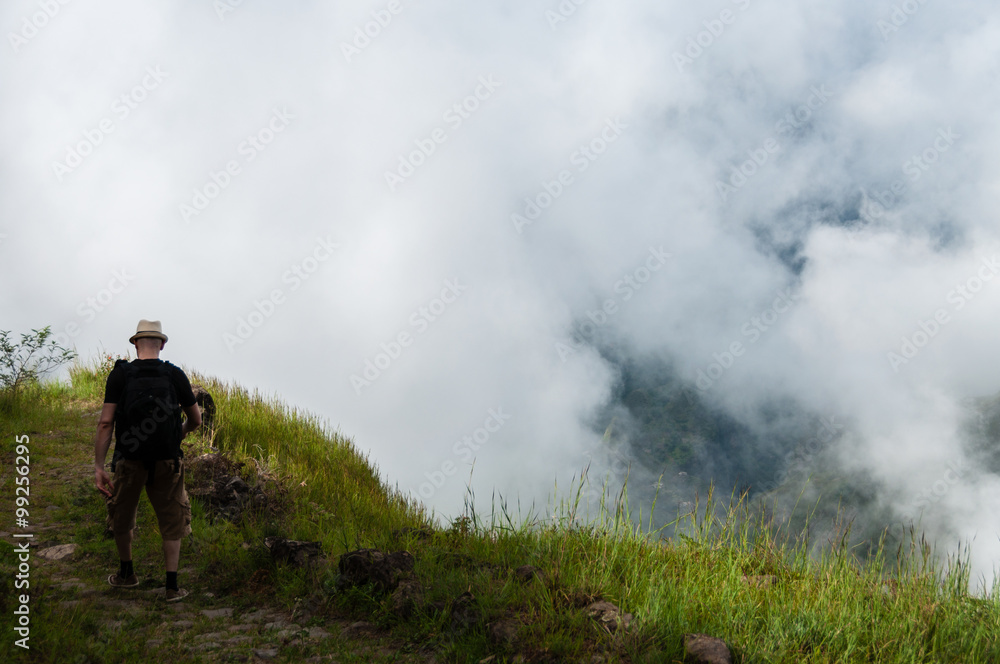 White man with black shirt and hat walking along a cliff above the clouds on mountain in cape verde