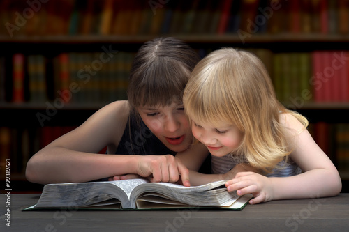 Дети читают книгу в библиотеке. Две сестры, девочки, читают книгу. Старшая сестра, подросток, водит пальцем по строчкам текста. Младшая девочка с интересом читает книгу