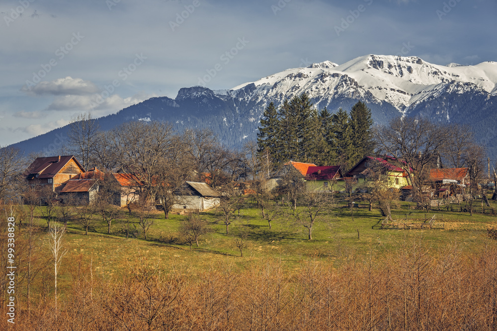 Spring sunny rural landscape with snowy Bucegi mountains and traditional Romanian houses and farm in Predelut village, near Bran, Transylvania region, Romania.