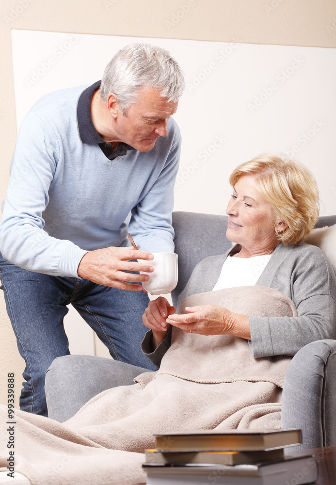 Obraz premium Portrait of lovely senior couple relaxing at home. Elderly woman sitting in armchair at living room with her husband. Old man holding in hand a glass of water while assisting his wife.