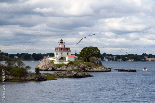 Sun illuminating Pomham Rocks lighthouse after stormy weather in Rhode Island as seagull flies in front of the beacon. Sun breaking through rain clouds.
