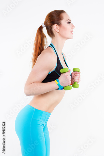 Young athletic red-haired woman in bright sportswear is doing exercise with small green dumbbells. She smiles brightly. © lina_aster