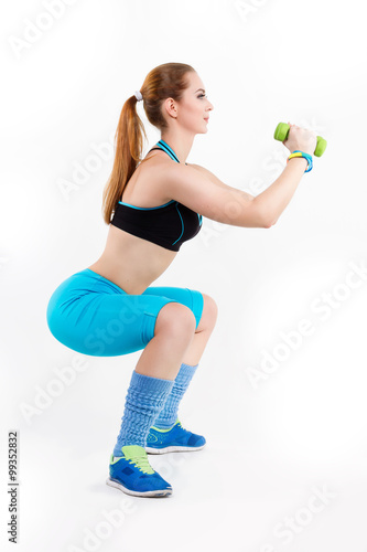 Young athletic red-haired woman in bright sportswear is doing squats with green dumbbells.