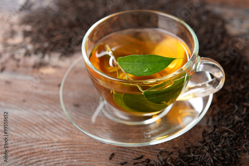 Glass cup of tea with green leaves on wooden background decorated with scattered tea