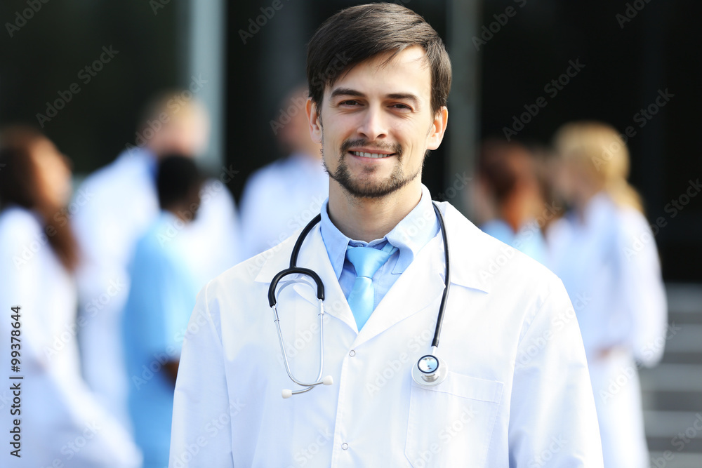 Smiling doctor with medical stuff behind standing against clinic entrance Smiling doctor with medical stuff behind standing against clinic entrance