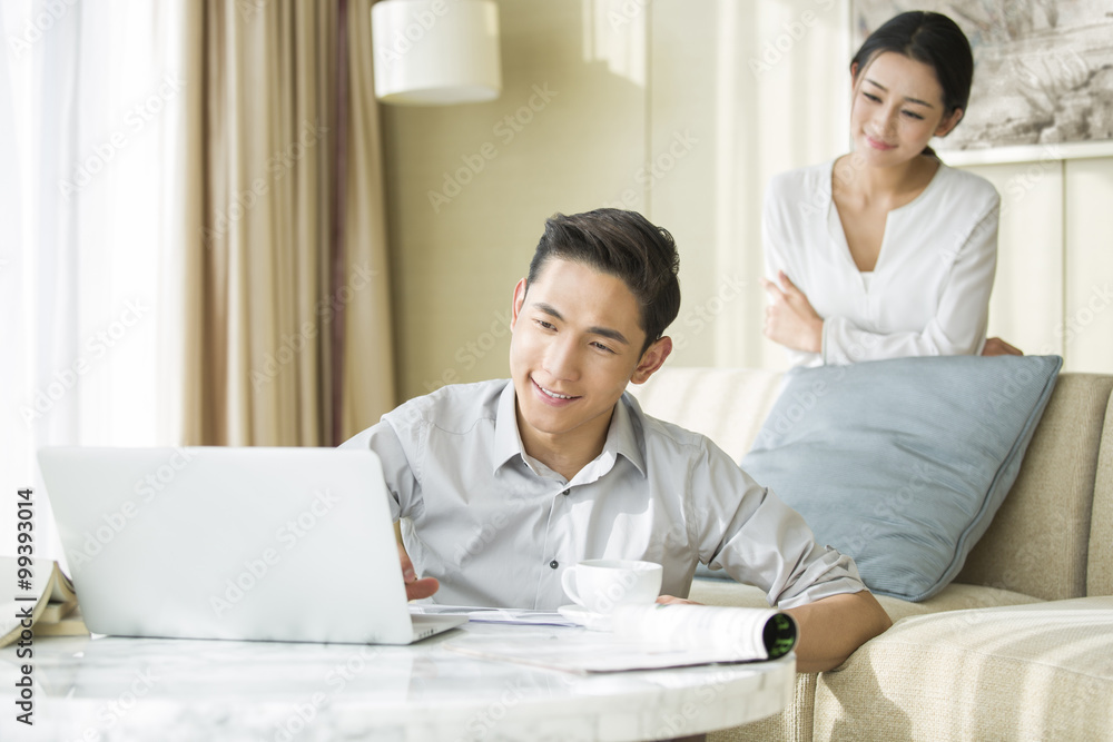 Young man working at home while his wife looking at him Young man working at home while his wife looking at him