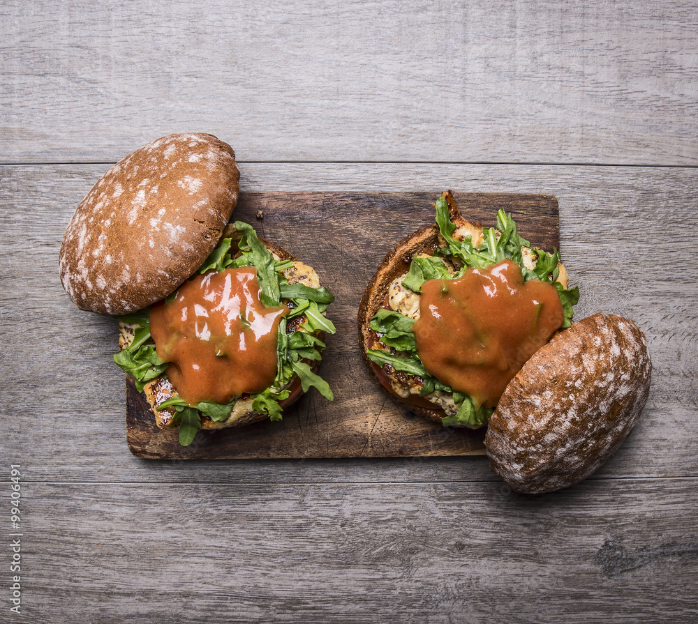 two home burgers with chicken in mustard sauce, arugula, tomatoes on a cutting board on wooden rustic background top view close up two home burgers with chicken in mustard sauce, arugula, tomatoes on a cutting board on wooden rustic background top view close up