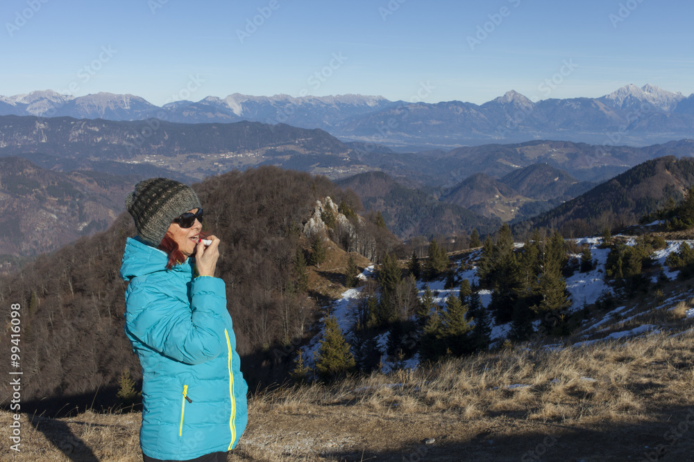 Naklejka premium Pretty mid aged woman trekker woman putting balsam lips to repair her lips of the causes of winter cold high in mountains on sunny winter day, space for text 