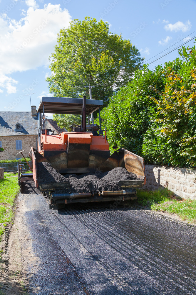 Road tarring machine using premix asphalt with the front flaps open waiting for a tip truck to reverse back in the narrow road refill the compartment Road tarring machine using premix asphalt with the front flaps open waiting for a tip truck to reverse back in the narrow road refill the compartment