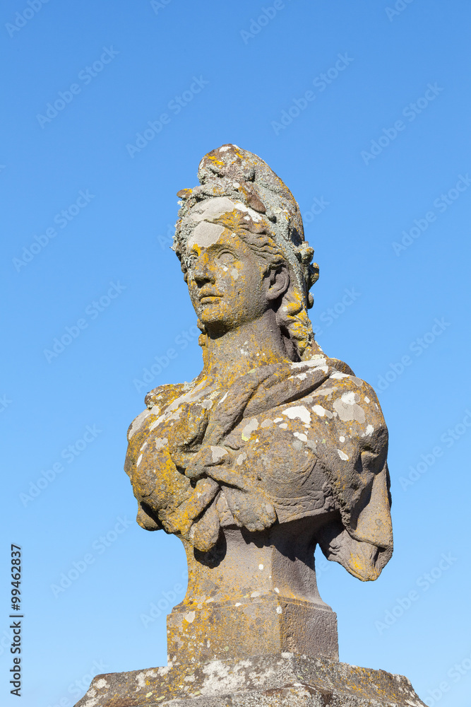 Od weathered stone bust of Marianne, a symbol of the French Republic and France, allegorical of Freedom and Reason, covered in colourful lichen against a clear blue sky with copy space