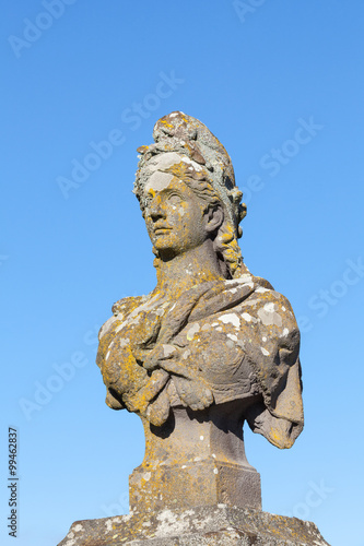 Od weathered stone bust of Marianne, a symbol of the French Republic and France, allegorical of Freedom and Reason, covered in colourful lichen against a clear blue sky with copy space