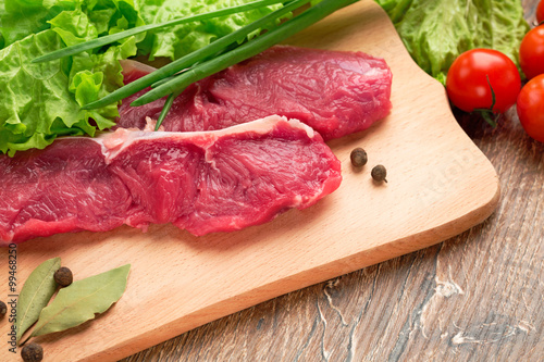 Fresh, crude stakes from meat veal on a wooden chopping board with setion,black pepper, lettuce leaves,fennel, bay leaf, green onions and cherry tomatoes